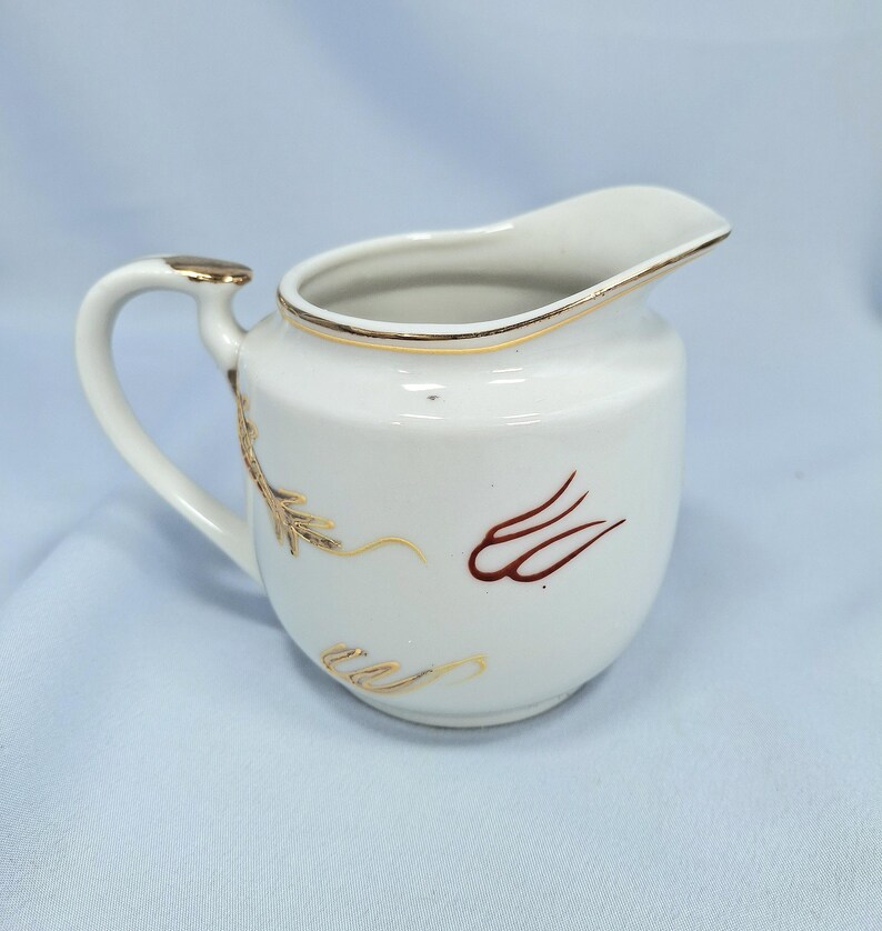 May include: White ceramic creamer with a curved handle and spout. The creamer has gold trim and decorative gold and brown leaf-like designs. The handle has a gold accent. The creamer is on a light blue surface.