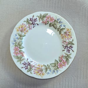 以下が含まれることがあります： 白いセラミックプレート、花柄のデザイン。プレートはスカラップエッジと金色の縁が特徴です。花柄には、ピンク、黄色、紫の花と緑の葉が含まれています。繊細でエレガントな作品です。