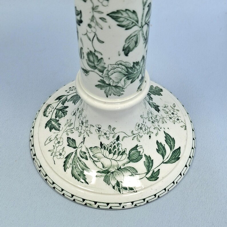 K&ouml;nnte beinhalten: Ein wei&szlig;er Kerzenst&auml;nder aus Keramik mit einem gr&uuml;nen Blumenmuster. Der Sockel ist rund mit einem detaillierten Rand, und der Stiel ist zylindrisch. Das Design zeigt Rosen und Bl&auml;tter und schafft einen klassischen, eleganten Look. Geeignet f&uuml;r die Inneneinrichtung.