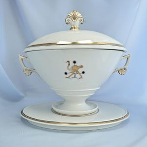 May include: White ceramic soup tureen with a lid, gold trim, and a decorative ostrich design. The tureen has handles and sits on a matching plate. The lid is topped with a gold finial.