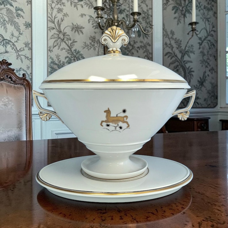 May include: A white porcelain serving dish with a lid, gold trim, and a gold deer emblem. The dish sits on a matching plate, and has ornate handles. The lid is topped with a decorative gold finial. The dish is on a wooden table.
