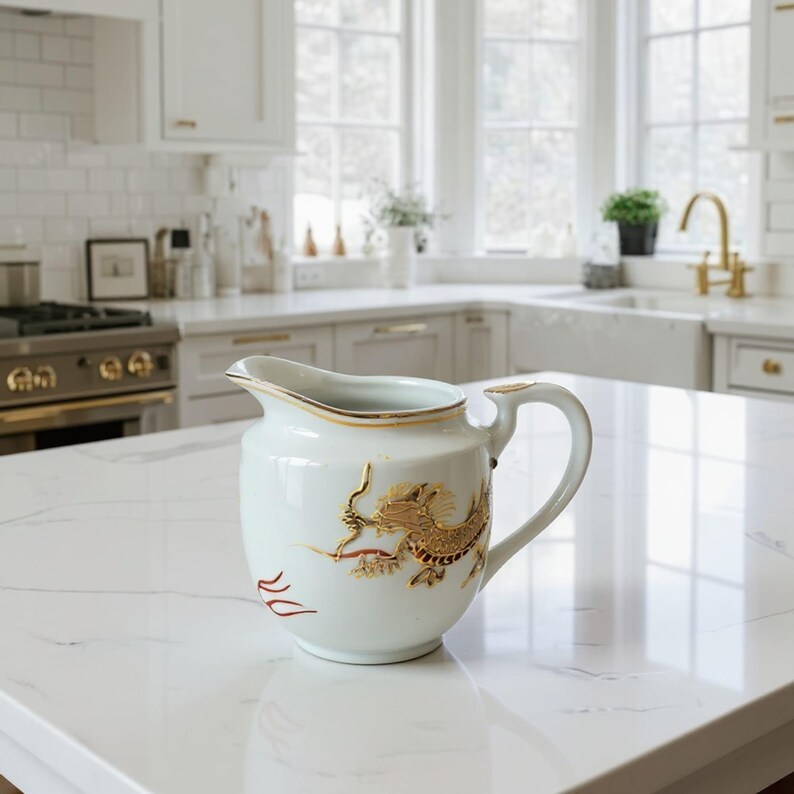 May include: A white ceramic jug with a gold rim and handle, decorated with a gold and red dragon motif. The jug is placed on a white countertop in a well-lit kitchen.