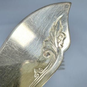 May include: Close-up of a silver serving utensil with a decorative floral design. The utensil has a curved, leaf-like shape with etched details. The silver surface shows signs of wear.