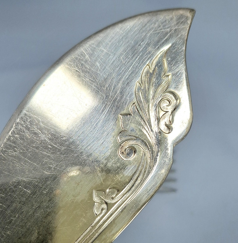 May include: Close-up of a silver serving utensil with a decorative floral design. The utensil has a curved, leaf-like shape with etched details. The silver surface shows signs of wear.