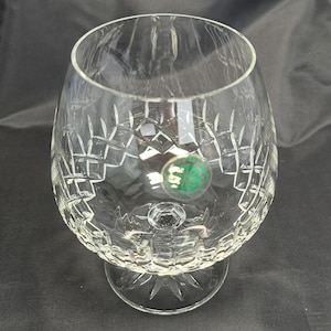 May include: A clear crystal brandy snifter featuring a wide, rounded bowl and a short, sturdy stem. The glass is decorated with intricate diamond-cut patterns and a starburst design on the base. A small, circular green sticker is attached to the side.