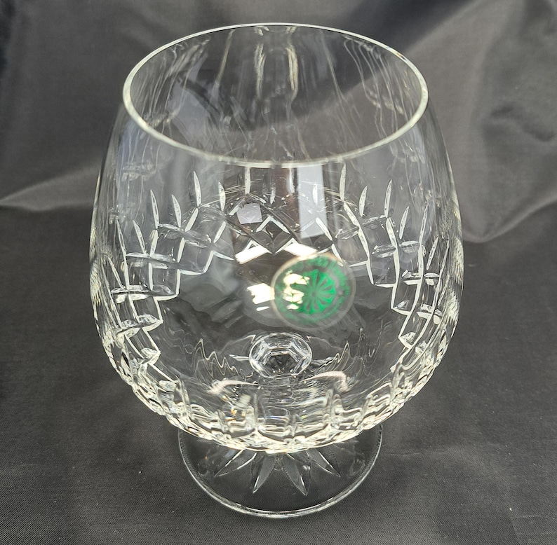 May include: A clear crystal brandy snifter featuring a wide, rounded bowl and a short, sturdy stem. The glass is decorated with intricate diamond-cut patterns and a starburst design on the base. A small, circular green sticker is attached to the side.