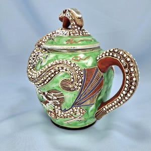 Pu&ograve; includere: Una teiera decorativa in ceramica verde con un design a drago. La teiera presenta un drago dettagliato avvolto attorno al corpo e al coperchio, con un manico e dettagli marroni. Il drago ha scaglie bianche e marroni e la teiera ha un bordo argentato.