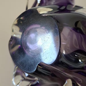 K&ouml;nnte beinhalten: Ein tiefviolettes Glasobjekt mit einem glatten, abgerundeten Boden in Form einer Blume mit f&uuml;nf ausgepr&auml;gten Lappen. Die Glasoberfl&auml;che weist sichtbare leichte Kratzer und Reflexionen auf und besitzt eine dunkle, durchscheinende Beschaffenheit.