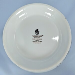 Puede incluir: Un plato de cer&aacute;mica blanca con un borde ligeramente curvado. El plato presenta el texto "WEDGWOOD Bone China MADE IN ENGLAND KUTANI CRANE &copy;WEDGWOOD 1971" en negro. Tambi&eacute;n hay un emblema decorativo en blanco y negro.