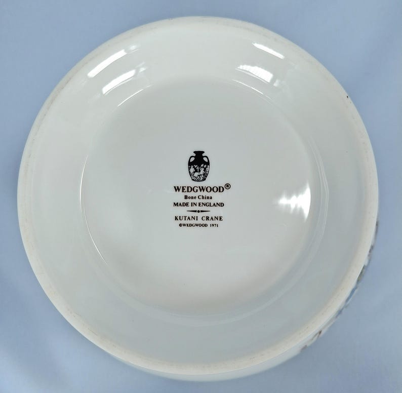 Puede incluir: Un plato de cer&aacute;mica blanca con un borde ligeramente curvado. El plato presenta el texto "WEDGWOOD Bone China MADE IN ENGLAND KUTANI CRANE &copy;WEDGWOOD 1971" en negro. Tambi&eacute;n hay un emblema decorativo en blanco y negro.