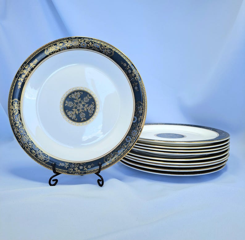 Op de afbeelding: Een set ronde witte keramische borden met een brede donkerblauwe rand met ingewikkelde gouden bloemmotieven en een bijpassend rond medaillon in goud en donkerblauw in het midden. E&eacute;n bord staat rechtop op een zwarte metalen standaard.