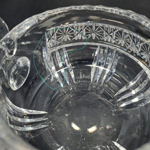 Op de afbeelding: Een heldere glazen kom met een decoratief geslepen patroon langs de bovenrand en verticale gegroefde inkepingen die naar de basis toe lopen. Een dunne, lichtblauwe cirkelvormige omtrek is zichtbaar op het binnenoppervlak van de kom.
