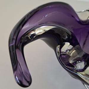 K&ouml;nnte beinhalten: Eine tiefviolette Glasschale mit einer flie&szlig;enden, organischen Form und dicken, geschwungenen R&auml;ndern. Das durchscheinende Material zeigt verschiedene Schattierungen von Violett und Dunkellila mit glatten, polierten Oberfl&auml;chen und Lichtreflexen.