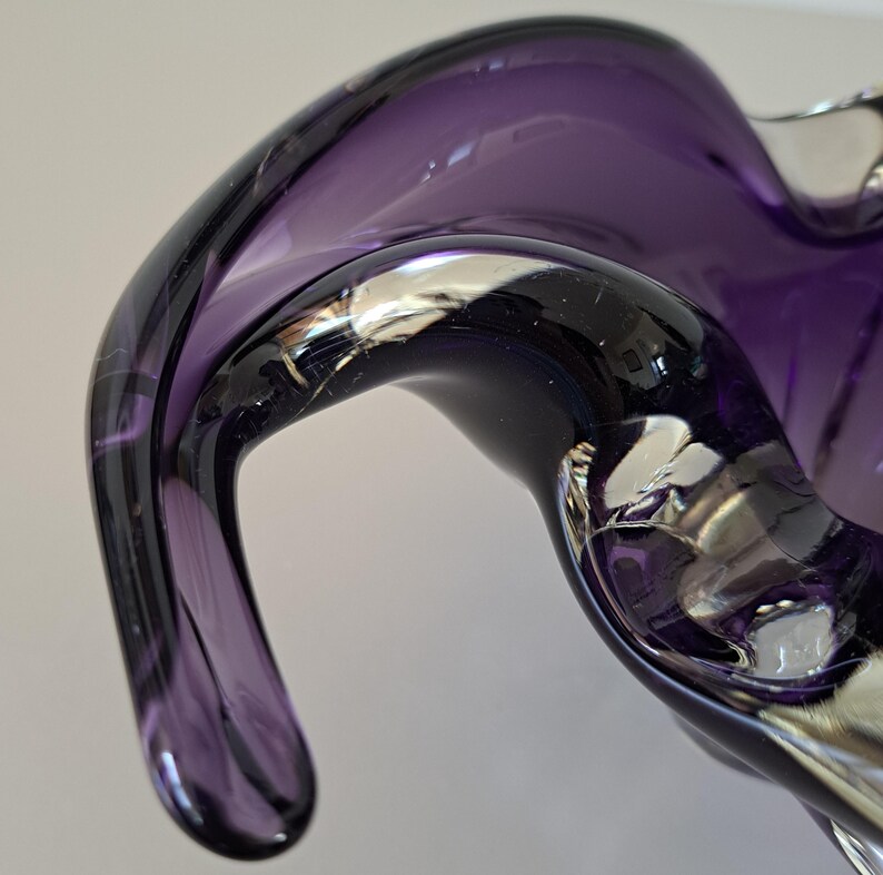 K&ouml;nnte beinhalten: Eine tiefviolette Glasschale mit einer flie&szlig;enden, organischen Form und dicken, geschwungenen R&auml;ndern. Das durchscheinende Material zeigt verschiedene Schattierungen von Violett und Dunkellila mit glatten, polierten Oberfl&auml;chen und Lichtreflexen.