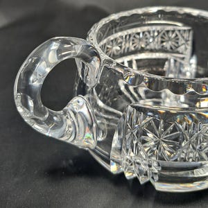 Op de afbeelding: Een creamer van helder glas met een gebogen handvat en ingewikkelde sterpatronen in het buitenoppervlak geslepen. De rand heeft een geschulpte afwerking en het lichaam vertoont geometrische facetten die het licht vangen en weerkaatsen.