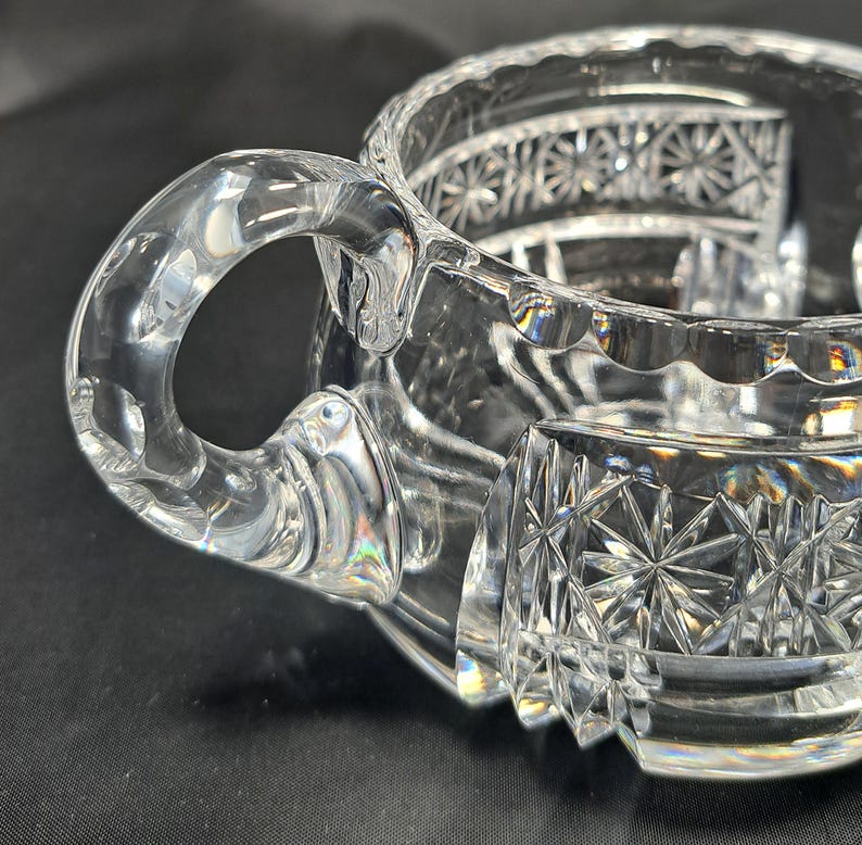 Op de afbeelding: Een creamer van helder glas met een gebogen handvat en ingewikkelde sterpatronen in het buitenoppervlak geslepen. De rand heeft een geschulpte afwerking en het lichaam vertoont geometrische facetten die het licht vangen en weerkaatsen.