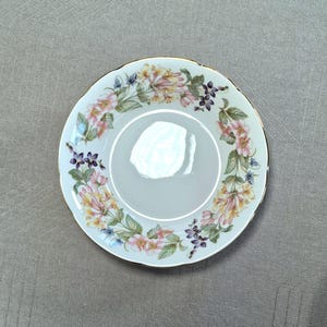 以下が含まれることがあります： 白いセラミックの受け皿で、花の模様が施されています。縁はスカラップで金色のトリムが施されています。花の模様には、ピンク、黄色、紫の花と緑の葉が円形に配置されています。