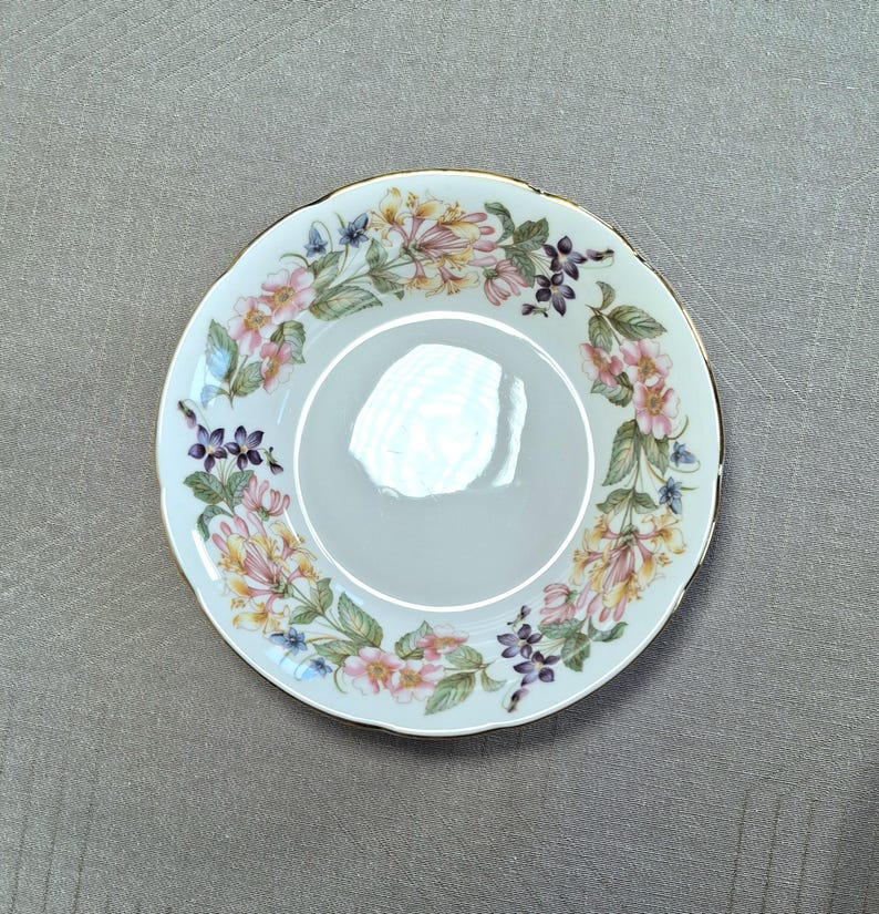 以下が含まれることがあります： 白いセラミックの受け皿で、花の模様が施されています。縁はスカラップで金色のトリムが施されています。花の模様には、ピンク、黄色、紫の花と緑の葉が円形に配置されています。