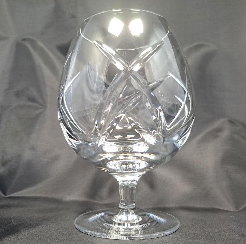 Puede incluir: Una copa de brandy de cristal transparente con un c&aacute;liz ancho y redondeado y un tallo corto y robusto. El vidrio est&aacute; decorado con profundos cortes geom&eacute;tricos angulares que crean un patr&oacute;n facetado en la superficie, reflejando la luz a trav&eacute;s del material.