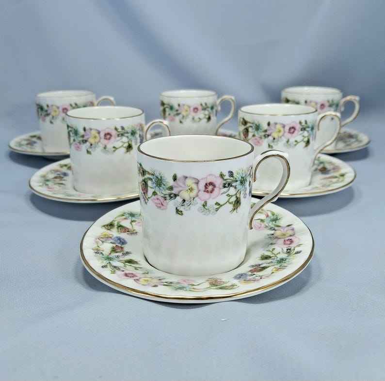 Peut inclure: Un ensemble de tasses et soucoupes en porcelaine blanche. Chaque tasse et soucoupe est orn&eacute;e d'un motif floral rose, jaune et bleu. Les bords des tasses et des soucoupes sont dor&eacute;s.