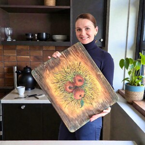 May include: A rectangular wooden chopping board with a scorched, dark-grained finish. The centre features a painted illustration of three red apples nestled in a patch of yellow and green grass, with a single green leaf visible at the bottom.
