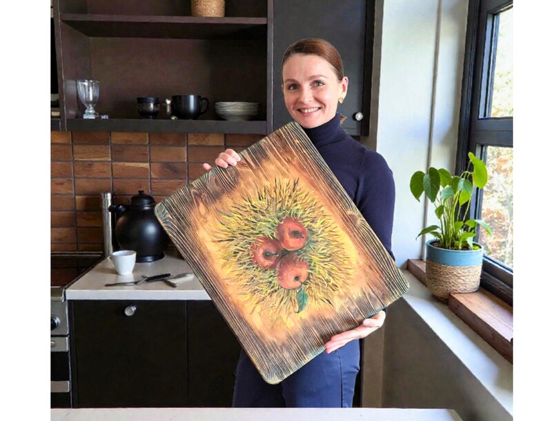 May include: A rectangular wooden chopping board with a scorched, dark-grained finish. The centre features a painted illustration of three red apples nestled in a patch of yellow and green grass, with a single green leaf visible at the bottom.