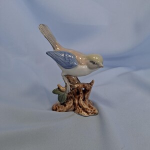 以下が含まれることがあります： 茶色の切り株に止まっているセラミック製の鳥の置物。鳥はベージュ色の体、青い翼、白い胸をしています。木の切り株は、小さな緑の葉が付いた、詳細で質感のある外観をしています。