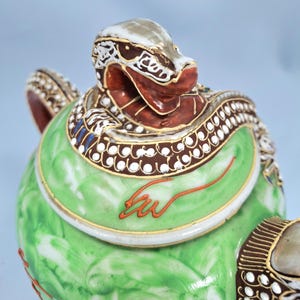 Pu&ograve; includere: Vista ravvicinata di un oggetto decorativo in ceramica. Presenta una testa a forma di serpente nei toni del marrone, bianco e rosso, su un corpo verde con dettagli dorati. Il design include motivi intricati e un disegno arancione dipinto a mano.