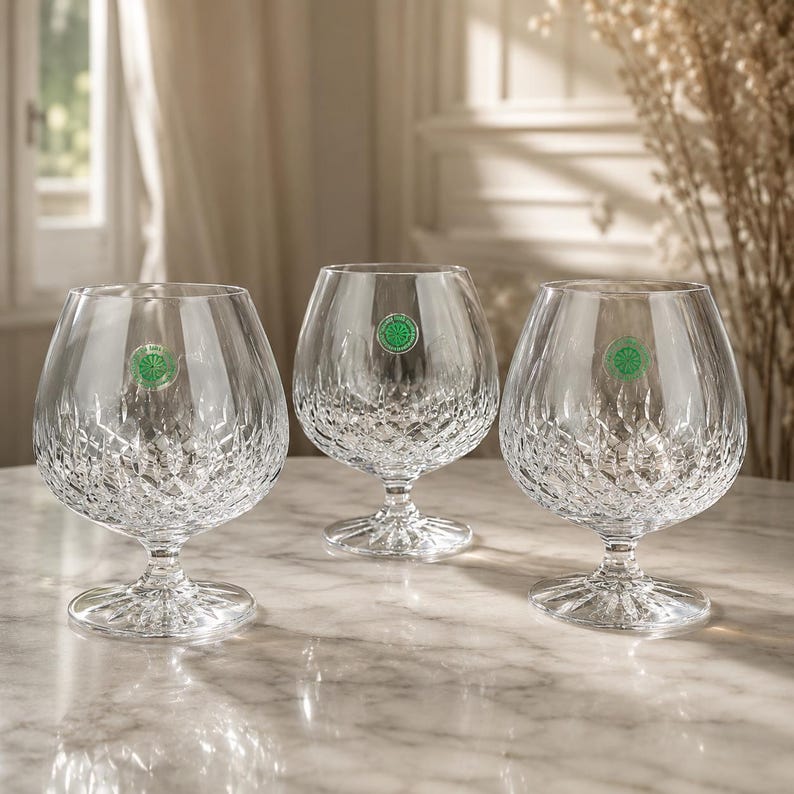 May include: Three clear crystal brandy snifters featuring a detailed diamond-cut pattern on the lower bowl and a starburst design on the circular base. Each glass has a small round green sticker with white text applied to the upper bowl.