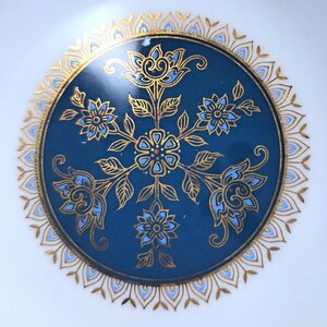 Op de afbeelding: Een rond keramieken bord met een diepblauw middenstuk, versierd met een symmetrisch goudkleurig en lichtblauw bloemmotief. Het ontwerp bevat een centrale bloem omringd door vier uitstralende bloesems, omlijst door een gedetailleerde gouden filigraanrand.