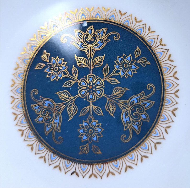 Op de afbeelding: Een rond keramieken bord met een diepblauw middenstuk, versierd met een symmetrisch goudkleurig en lichtblauw bloemmotief. Het ontwerp bevat een centrale bloem omringd door vier uitstralende bloesems, omlijst door een gedetailleerde gouden filigraanrand.