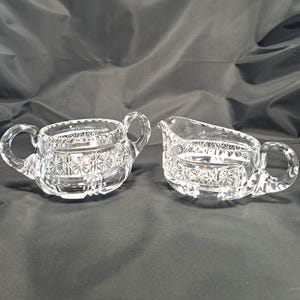 Op de afbeelding: Een suikerpot van helder glas met twee zijhandvatten en een bijpassende melkkan, beide voorzien van een geperst sterpatroon en horizontale banden. De set is gemaakt van transparant glas met een gefacetteerd ontwerp en een gepolijste afwerking.