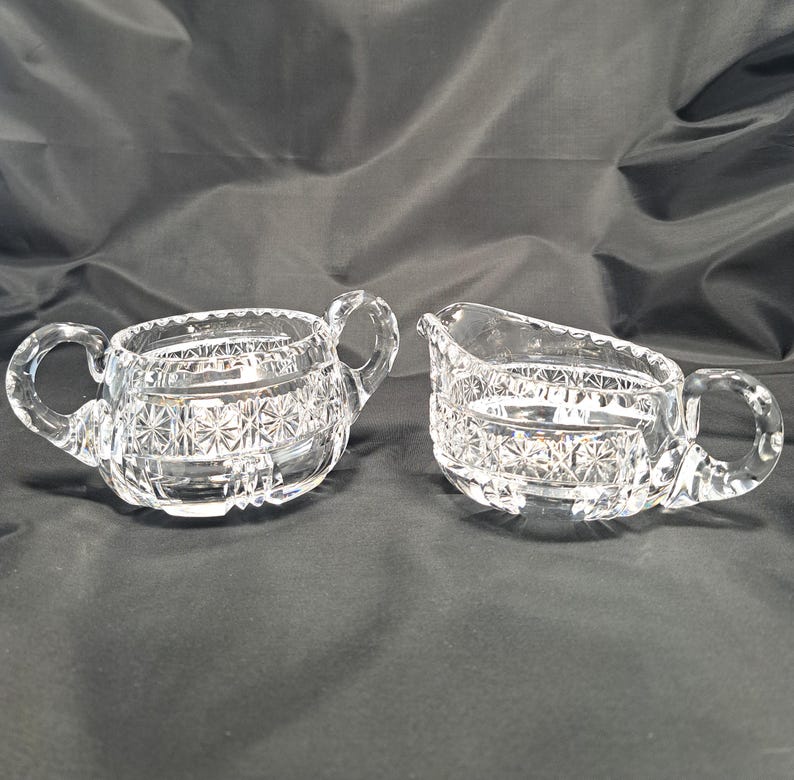 Op de afbeelding: Een suikerpot van helder glas met twee zijhandvatten en een bijpassende melkkan, beide voorzien van een geperst sterpatroon en horizontale banden. De set is gemaakt van transparant glas met een gefacetteerd ontwerp en een gepolijste afwerking.
