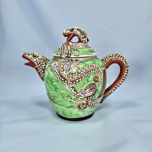 Pu&ograve; includere: Una teiera in ceramica con un design a drago. La teiera &egrave; principalmente verde con dettagli marroni e bianchi. Il manico e il beccuccio sono marroni e il coperchio presenta una testa di drago. Il design del drago avvolge il corpo della teiera.