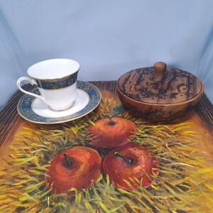 May include: A white porcelain teacup and saucer with a blue and gold patterned rim, placed next to a round wooden lidded bowl with dark grain patterns, all resting on a painted surface featuring three red apples nestled in yellow straw.