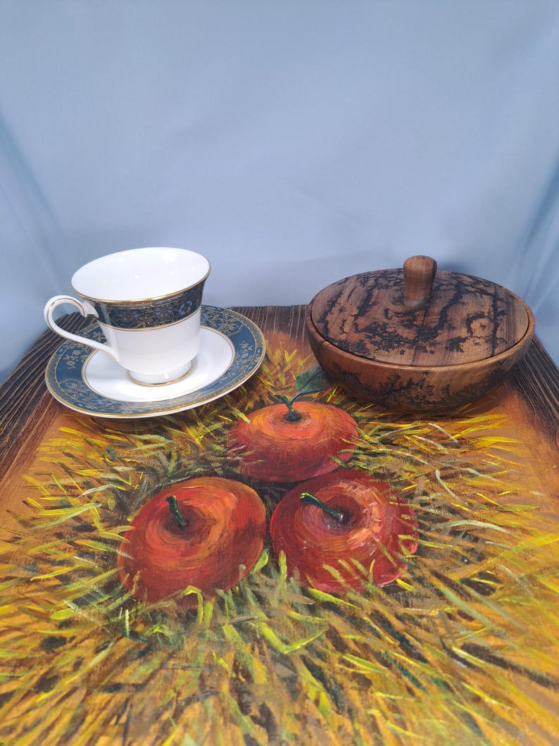 May include: A white porcelain teacup and saucer with a blue and gold patterned rim, placed next to a round wooden lidded bowl with dark grain patterns, all resting on a painted surface featuring three red apples nestled in yellow straw.