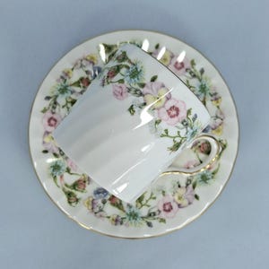 Peut inclure: Service &agrave; th&eacute; blanc avec une tasse et une soucoupe orn&eacute;es de motifs floraux roses, jaunes et bleus. La tasse a une anse dor&eacute;e et la soucoupe est festonn&eacute;e. L'ensemble est probablement en porcelaine.