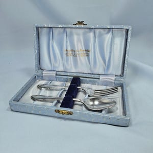 May include: A vintage cutlery set in a light blue box. The box is lined with light blue satin and holds a fork, spoon, and knife. The box has the text "Eucley & Moorite Cutlers & Silversmiths Sheffield - England".