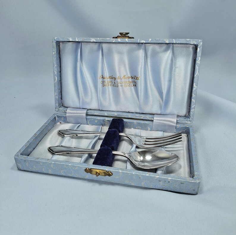 May include: A vintage cutlery set in a light blue box. The box is lined with light blue satin and holds a fork, spoon, and knife. The box has the text "Eucley & Moorite Cutlers & Silversmiths Sheffield - England".