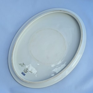 May include: White oval porcelain serving platter. The underside of the platter features a faded circular design and a blue and white emblem with text. The platter has a slightly raised edge.