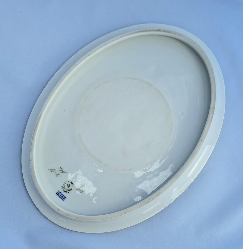 May include: White oval porcelain serving platter. The underside of the platter features a faded circular design and a blue and white emblem with text. The platter has a slightly raised edge.