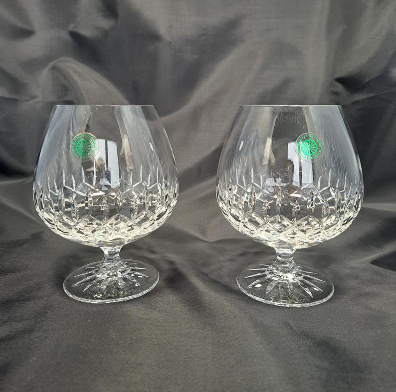 May include: A pair of clear crystal brandy snifters featuring a deep diamond-cut pattern on the lower bowl and a starburst design on the circular base. Each glass has a small circular green sticker applied to the upper side of the bowl.