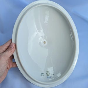 May include: An oval, white ceramic serving dish with a slightly raised rim. The dish has a small, circular indentation in the center and a small blue stamp with text on the bottom. The dish is held by a hand.