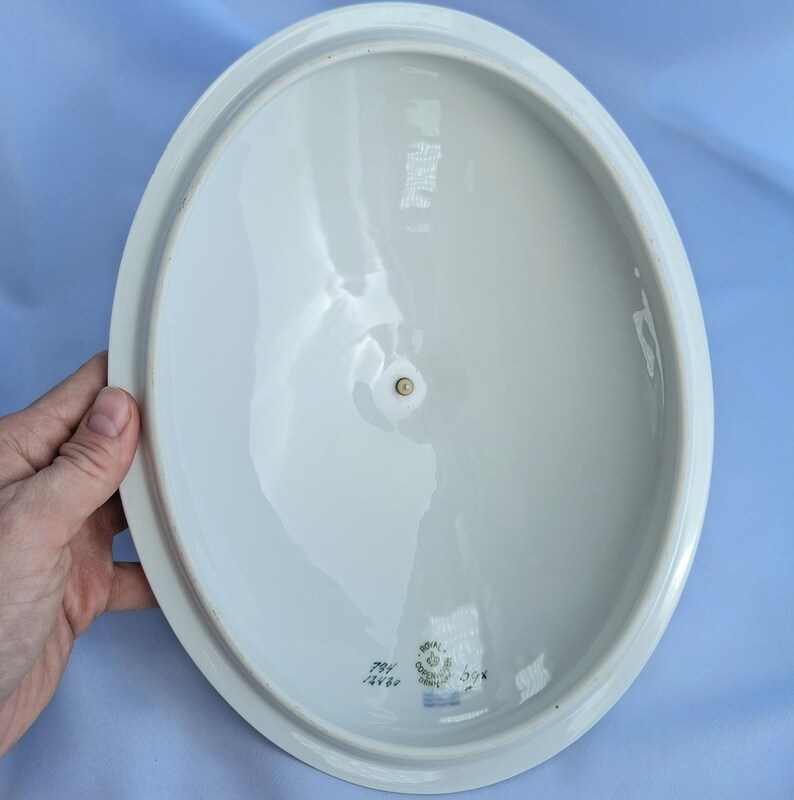 May include: An oval, white ceramic serving dish with a slightly raised rim. The dish has a small, circular indentation in the center and a small blue stamp with text on the bottom. The dish is held by a hand.