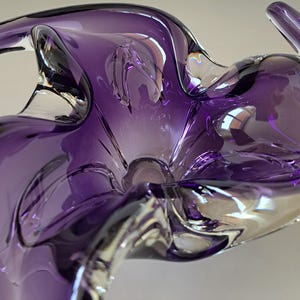 K&ouml;nnte beinhalten: Eine tiefviolette Glasschale mit einer freien, organischen Form und gewellten R&auml;ndern. Das durchscheinende Glas geht von intensiven Violettt&ouml;nen in klare Bereiche &uuml;ber und erzeugt ein dynamisches, flie&szlig;endes Erscheinungsbild mit Lichtreflexen.