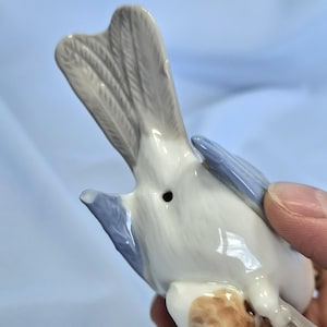 以下が含まれることがあります： 詳細な尾と翼を持つ磁器製の鳥の置物。鳥は白、水色、グレーの色を組み合わせています。置物は人の指で持たれています。