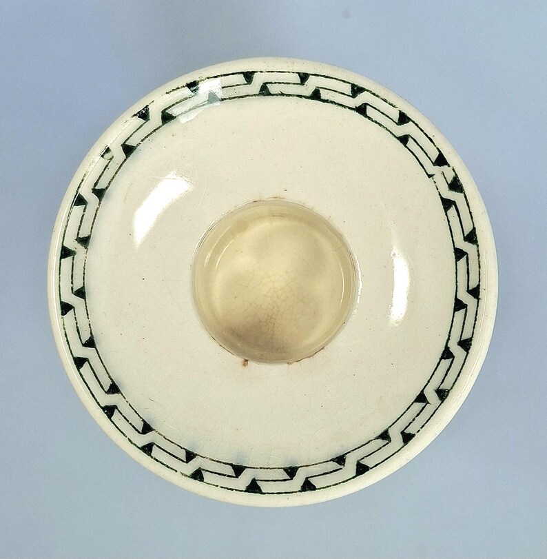 K&ouml;nnte beinhalten: Eine wei&szlig;e Keramikschale mit einem zentralen, vertieften Kreis. Der Rand ist mit einem sich wiederholenden schwarzen geometrischen Muster verziert. Die Schale hat eine gl&auml;nzende Oberfl&auml;che und wird von oben betrachtet.