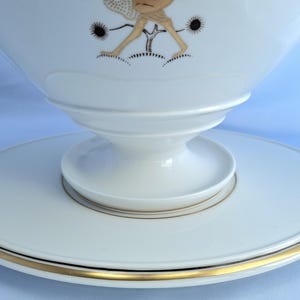 May include: White ceramic serving bowl with a pedestal base and a wide, flat rim. The bowl features a gold and black illustration of a figure. The rim is accented with a thin gold band. The bowl is likely for serving food.
