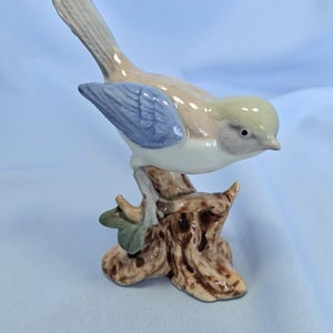 以下が含まれることがあります： 茶色の切り株に止まっているセラミック製の鳥の置物。鳥は薄茶色の体、青い翼、黄色の頭をしています。この置物は光沢のある仕上げと小さな緑の葉で詳細に表現されています。