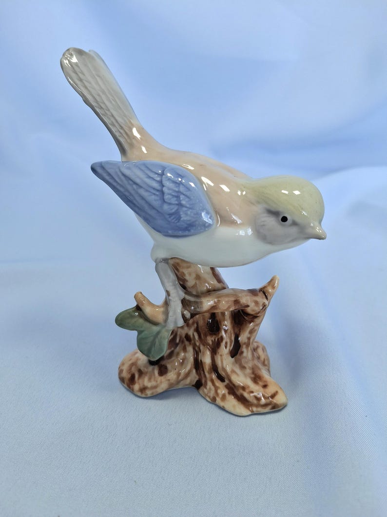 以下が含まれることがあります： 茶色の切り株に止まっているセラミック製の鳥の置物。鳥は薄茶色の体、青い翼、黄色の頭をしています。この置物は光沢のある仕上げと小さな緑の葉で詳細に表現されています。